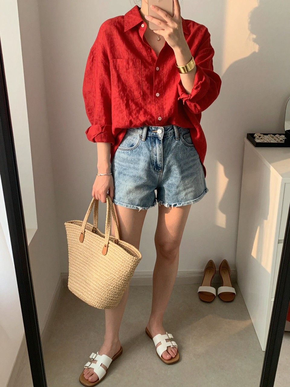 Image Prompt:
mirror selfie of a woman wearing a red oversized linen button-down shirt, light-wash denim cutoff shorts, white leather slide sandals, a natural straw tote bag, and a gold cuff bracelet, clearly showing the same clothing items, colors, fabrics, and accessories listed in the Outfit Pieces section, holding a phone in front of a mirror, cropped at NECK so the face does not show, with a relaxed casual selfie pose, in a fashion studio mirror corner, evening golden hour, with a pair of heels on the floor nearby and a jewelry tray on the vanity, natural casual mirror selfie, not staged, latest iPhone photo quality.