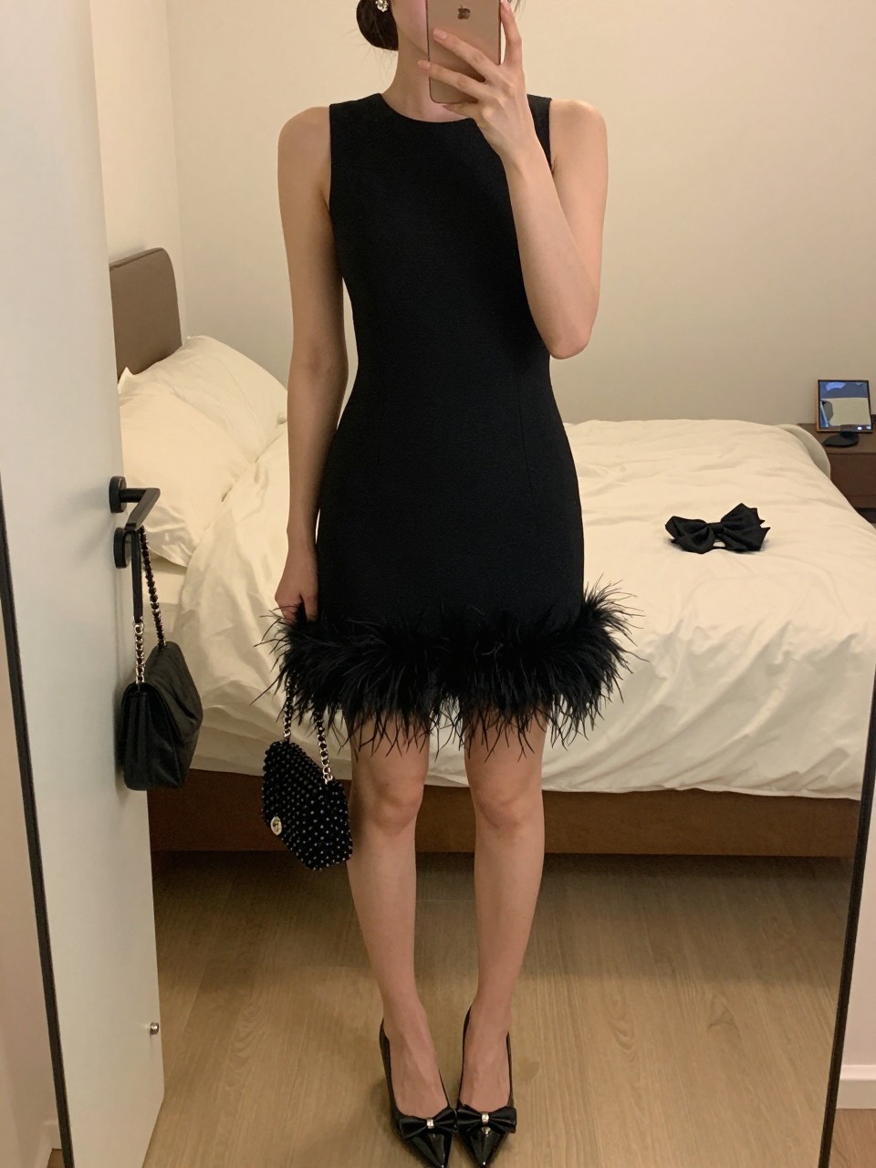 mirror selfie of a woman wearing a black cocktail mini dress with a feather-trim hem, black patent bow pumps, crystal stud earrings, a small black beaded bag, and a polished low bun with soft shimmer eyeshadow, clearly showing the same clothing items, colors, fabrics, and accessories listed in the Outfit Pieces section, holding a phone in front of a mirror, cropped at NECK so the face does not show, with a relaxed casual selfie pose, in a feminine-themed bedroom, evening golden hour, including soft pillows on an unmade bed and a handbag hanging on a door handle, latest iPhone photo quality.