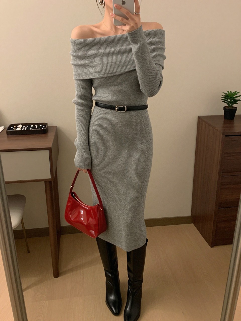 Outfit Image Prompt:
mirror selfie of a woman wearing a gray off-shoulder ribbed knit midi dress, black knee-high boots, red patent leather mini bag, silver hoop earrings, and a slim black belt at the waist, holding a phone in front of a mirror, cropped at NECK so the face does not show, relaxed high-fashion mirror selfie pose, in a minimal dressing room, warm moody lighting, with a jewelry tray on the vanity and a small plant on a dresser, realistic latest iPhone photo quality, polished city outfit.