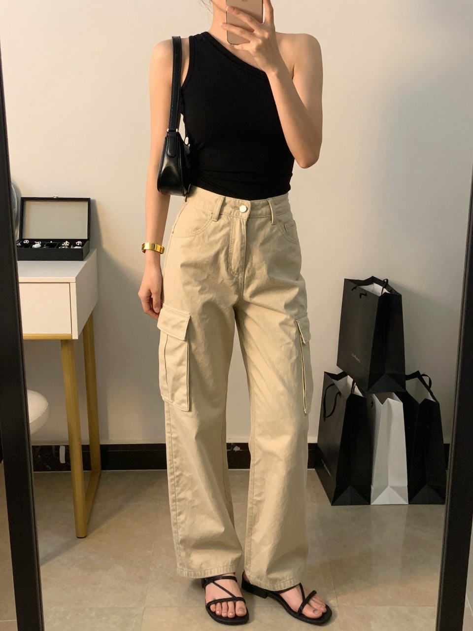 mirror selfie of a woman wearing beige cargo jeans, fitted black one-shoulder top, black strappy flat sandals, black mini shoulder bag, and gold cuff bracelet, clearly showing the same clothing items, colors, fabrics, and accessories listed in the Outfit Pieces section, holding a phone in front of a mirror, cropped at NECK so the face does not show, with a relaxed casual selfie pose, in a minimal dressing room, evening golden hour, including a jewelry tray on the vanity and shopping bags beside a chair, latest iPhone photo quality.