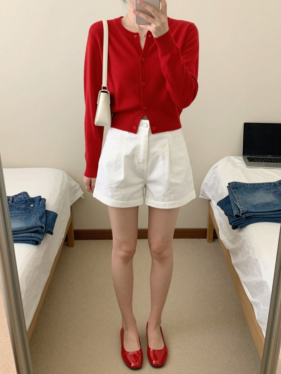 mirror selfie of a woman wearing a red cropped cardigan buttoned at the front, white high-waisted cotton shorts, glossy red jelly shoes, white mini shoulder bag, and delicate gold necklace, clearly showing the same clothing items, colors, fabrics, and accessories listed in the Outfit Pieces section, holding a phone in front of a mirror, cropped at NECK so the face does not show, with a relaxed casual selfie pose, in a minimal dressing room, soft indoor lighting, including a laptop on the bed and folded denim jeans on a bench, latest iPhone photo quality.