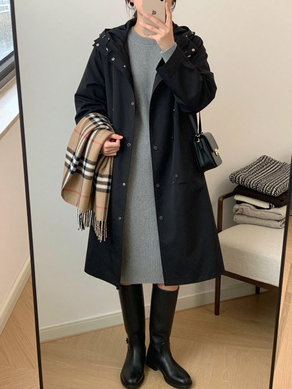 mirror selfie of a woman wearing the exact outfit described above, clearly showing a black hooded raincoat layered over a gray ribbed sweater midi dress, black knee-high rain boots, a black mini shoulder bag worn close to the body, a folded plaid scarf, and silver hoop earrings, holding a phone in front of a mirror, cropped at NECK so the face does not show, with a relaxed casual selfie pose, in a modern bedroom mirror setup, soft indoor lighting, with rain visible on the window and a folded blanket on a chair, latest iPhone photo quality.