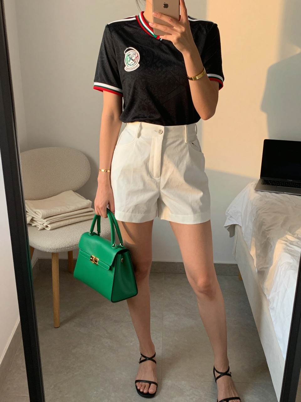 mirror selfie of a woman wearing a black Mexico soccer jersey tucked into white tailored shorts, black strappy flat sandals, green structured mini bag, and gold cuff bracelet, clearly showing the same clothing items, colors, fabrics, and accessories listed in the Outfit Pieces section, holding a phone in front of a mirror, cropped at NECK so the face does not show, with a relaxed casual selfie pose, in a minimal dressing room, evening golden hour, including a folded blanket on a chair and a laptop on the bed, latest iPhone photo quality.