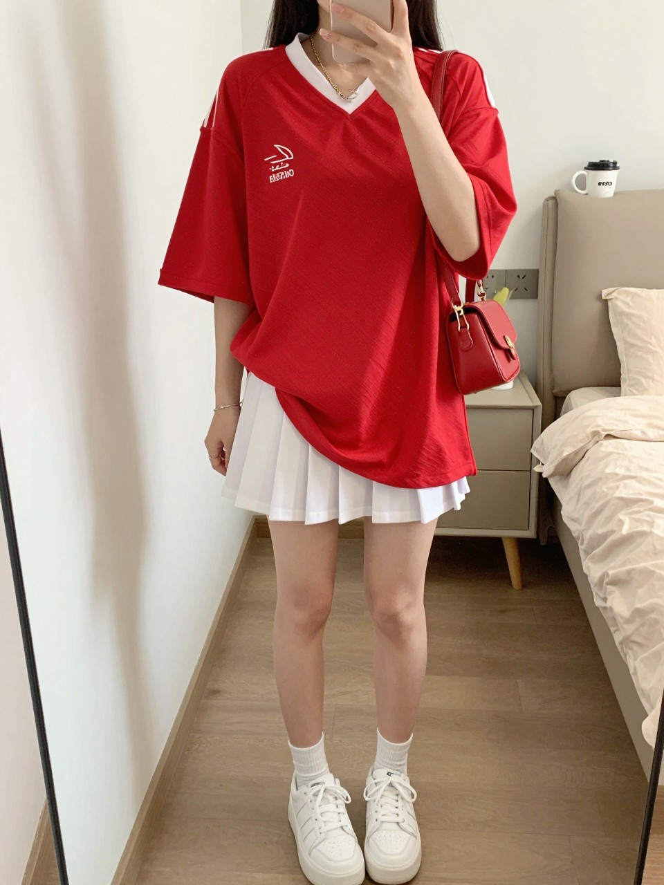 mirror selfie of a woman wearing an oversized red soccer jersey, white pleated tennis-style mini skirt, white platform sneakers, red mini shoulder bag, and slim gold chain necklace, clearly showing the same clothing items, colors, fabrics, and accessories listed in the Outfit Pieces section, holding a phone in front of a mirror, cropped at NECK so the face does not show, with a relaxed casual selfie pose, in a feminine-themed bedroom, natural window light, including a handbag hanging on a door handle and a coffee mug on a bedside table, latest iPhone photo quality.