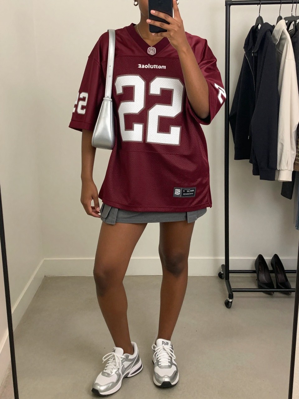 mirror selfie of a Black woman wearing an oversized burgundy football jersey, gray cargo mini skirt, white and silver sneakers, silver shoulder bag, and small silver huggie earrings, clearly showing the same clothing items, colors, fabrics, and accessories listed in the Outfit Pieces section, holding a phone in front of a mirror, cropped at NECK so the face does not show, with a relaxed casual selfie pose, in a minimal dressing room, soft indoor lighting, including a clothing rack with garments behind and a pair of heels on the floor nearby, latest iPhone photo quality.
