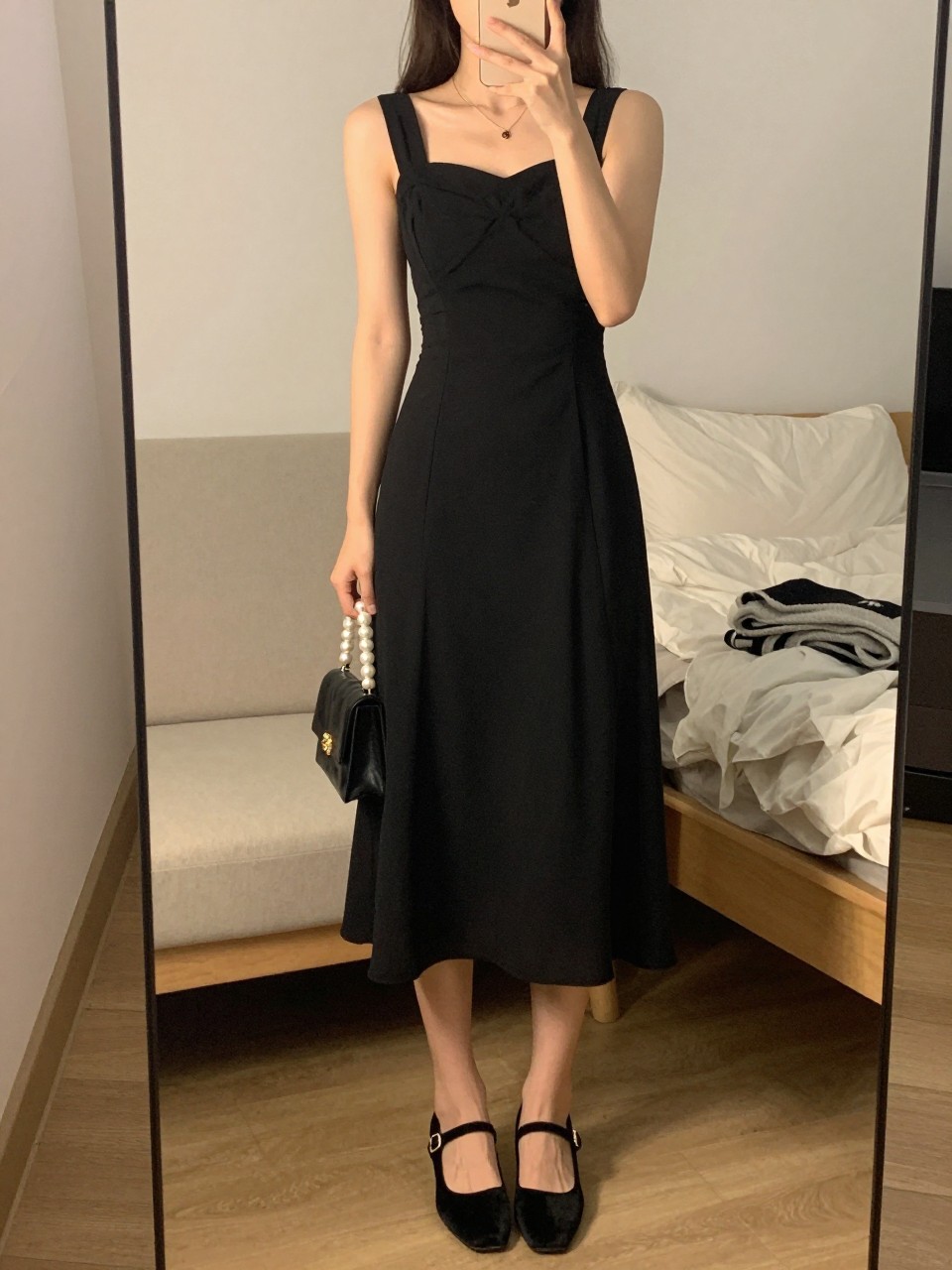 mirror selfie of a woman wearing a black pleated chiffon midi cocktail dress, black velvet Mary Jane heels, dainty gold layered necklace, a black pearl-handle bag, and a soft half-up hairstyle with peachy makeup, clearly showing the same clothing items, colors, fabrics, and accessories listed in the Outfit Pieces section, holding a phone in front of a mirror, cropped at NECK so the face does not show, with a relaxed casual selfie pose, in a cozy bedroom mirror setup, evening golden hour, including soft pillows on an unmade bed and a folded blanket on a chair, latest iPhone photo quality.