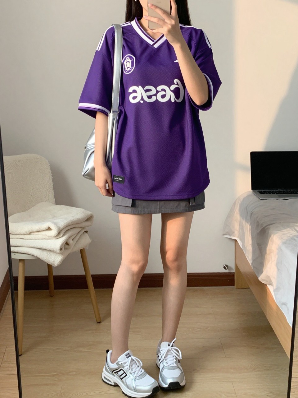 mirror selfie of a woman wearing an oversized purple soccer jersey, gray cargo mini skirt, white and silver sneakers, silver shoulder bag, and small silver huggie earrings, clearly showing the same clothing items, colors, fabrics, and accessories listed in the Outfit Pieces section, holding a phone in front of a mirror, cropped at NECK so the face does not show, with a relaxed casual selfie pose, in a minimal dressing room, evening golden hour, including a folded blanket on a chair and a laptop on the bed, latest iPhone photo quality.