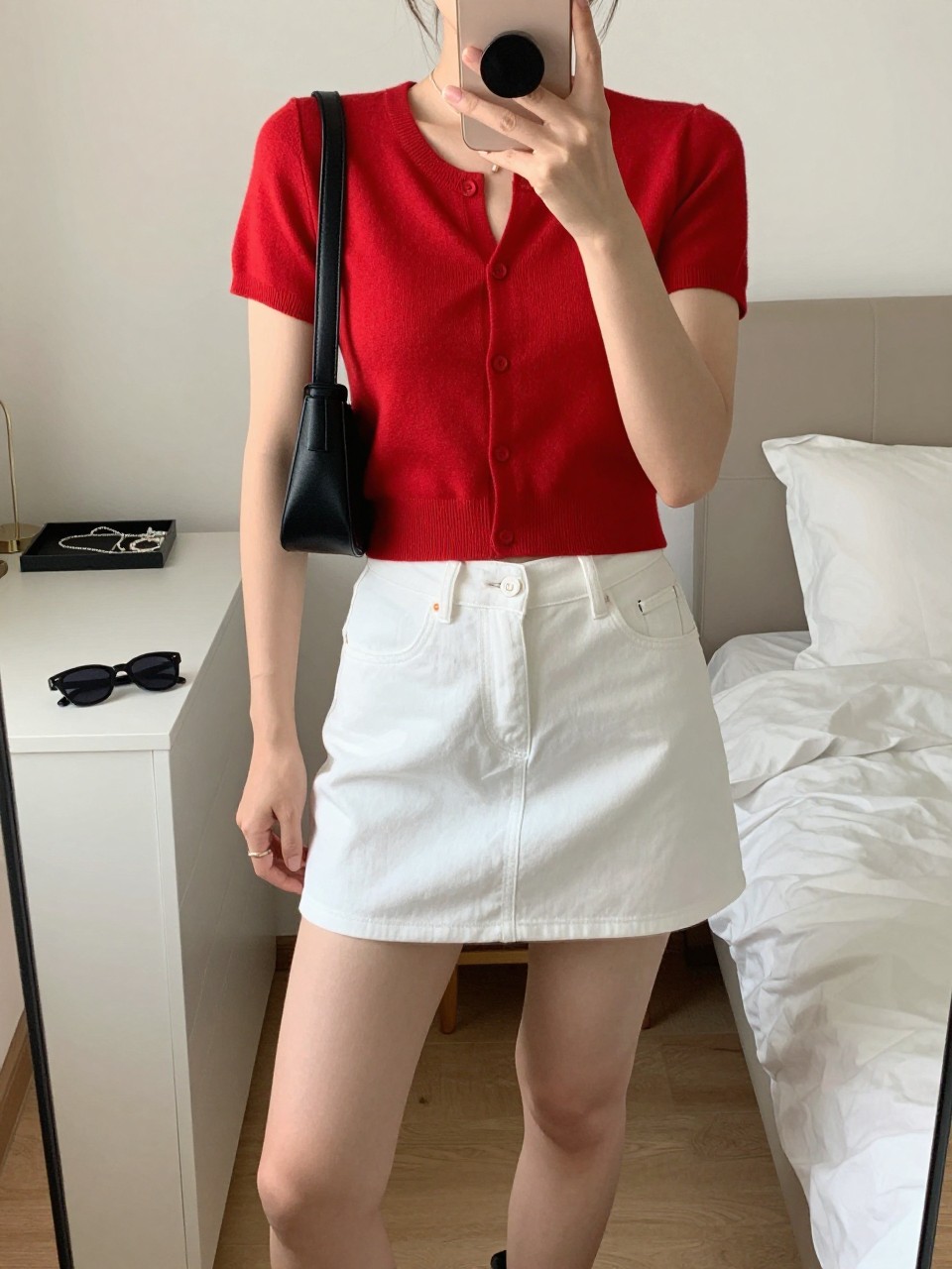 Outfit Image Prompt:
mirror selfie of a woman wearing a red short-sleeve cropped cardigan, white denim mini skirt, black platform sandals, small black leather shoulder bag, and slim black sunglasses, holding a phone in front of a mirror, cropped at NECK so the face does not show, relaxed casual selfie pose, in a feminine-themed bedroom, soft indoor lighting, with a jewelry tray on the vanity and sunglasses placed on a dresser, realistic latest iPhone photo quality, cute NYC summer outfit.