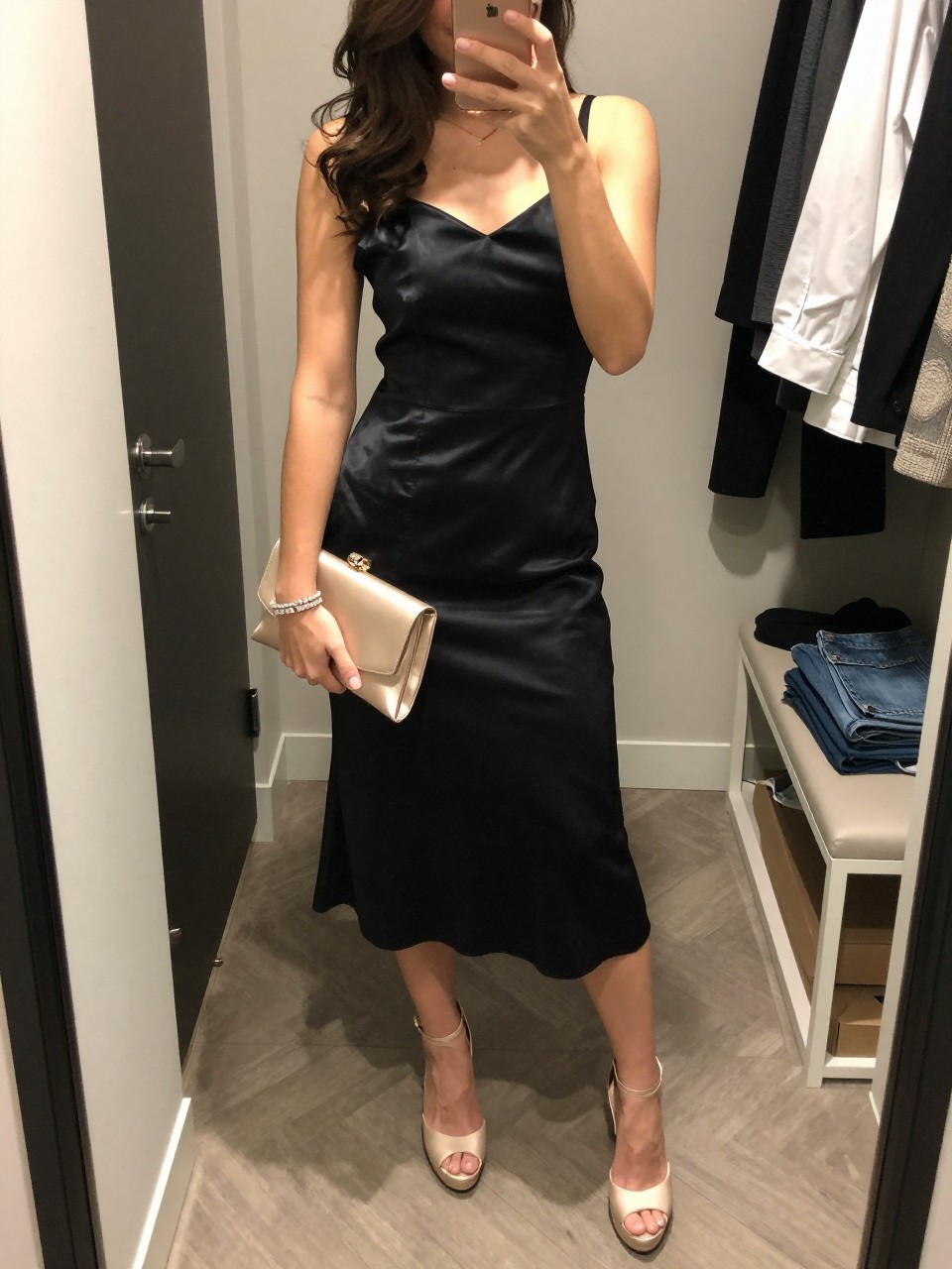 mirror selfie of a woman wearing a black liquid satin A-line midi cocktail dress, champagne platform heels, crystal tennis bracelet, a champagne satin clutch, and smooth waves with soft champagne eyeshadow, clearly showing the same clothing items, colors, fabrics, and accessories listed in the Outfit Pieces section, holding a phone in front of a mirror, cropped at NECK so the face does not show, with a relaxed casual selfie pose, in a walk-in closet mirror, soft indoor lighting, including open closet with clothes visible and folded denim jeans on a bench, latest iPhone photo quality.