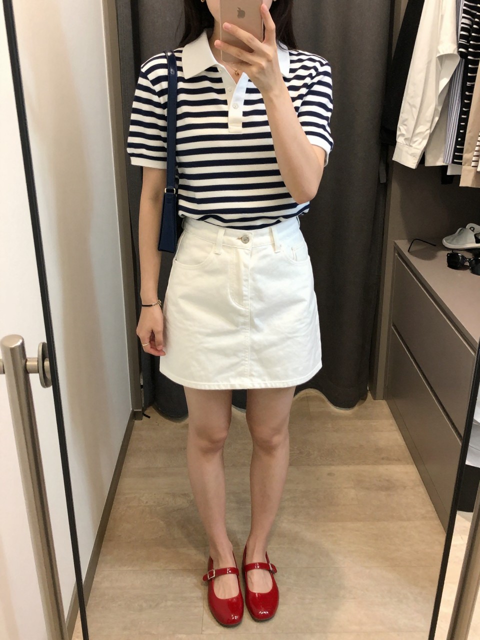 mirror selfie of a woman wearing a navy and white striped polo shirt tucked into white denim mini skirt, glossy red jelly shoes, navy mini shoulder bag, and small gold hoop earrings, clearly showing the same clothing items, colors, fabrics, and accessories listed in the Outfit Pieces section, holding a phone in front of a mirror, cropped at NECK so the face does not show, with a relaxed casual selfie pose, in a walk-in closet mirror, soft indoor lighting, including an open closet with clothes visible and sunglasses placed on a dresser, latest iPhone photo quality.