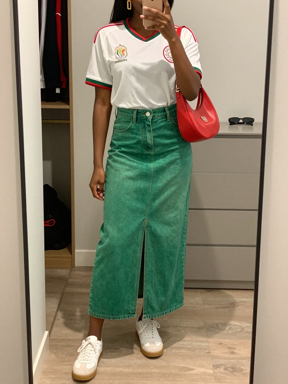 mirror selfie of a Black woman wearing a white Mexico soccer jersey, green denim maxi skirt with front slit, white leather sneakers, red crescent shoulder bag, and gold drop earrings, clearly showing the same clothing items, colors, fabrics, and accessories listed in the Outfit Pieces section, holding a phone in front of a mirror, cropped at NECK so the face does not show, with a relaxed casual selfie pose, in a walk-in closet mirror, soft indoor lighting, including an open closet with clothes visible and sunglasses placed on a dresser, latest iPhone photo quality.