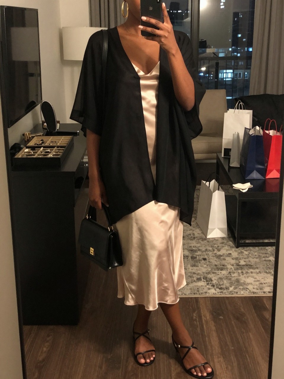 mirror selfie of a Black woman wearing a black sheer poncho layered over a champagne satin slip dress, black strappy flat sandals, black mini shoulder bag, and chunky gold hoop earrings, clearly showing the same clothing items, colors, fabrics, and accessories listed in the Outfit Pieces section, holding a phone in front of a mirror, cropped at NECK so the face does not show, with a relaxed casual selfie pose, in a stylish city apartment, warm moody lighting, including a jewelry tray on the vanity and shopping bags beside a chair, latest iPhone photo quality.