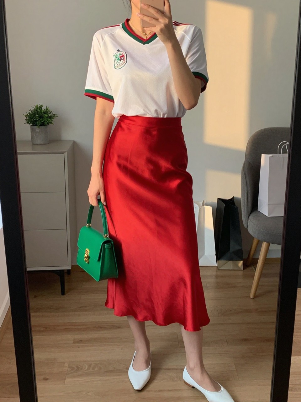 mirror selfie of a woman wearing a white Mexico soccer jersey tucked into a red satin midi skirt, white pointed ballet flats, green mini handbag, and delicate gold necklace, clearly showing the same clothing items, colors, fabrics, and accessories listed in the Outfit Pieces section, holding a phone in front of a mirror, cropped at NECK so the face does not show, with a relaxed casual selfie pose, in a stylish city apartment, evening golden hour, including a small plant on a dresser and shopping bags beside a chair, latest iPhone photo quality.