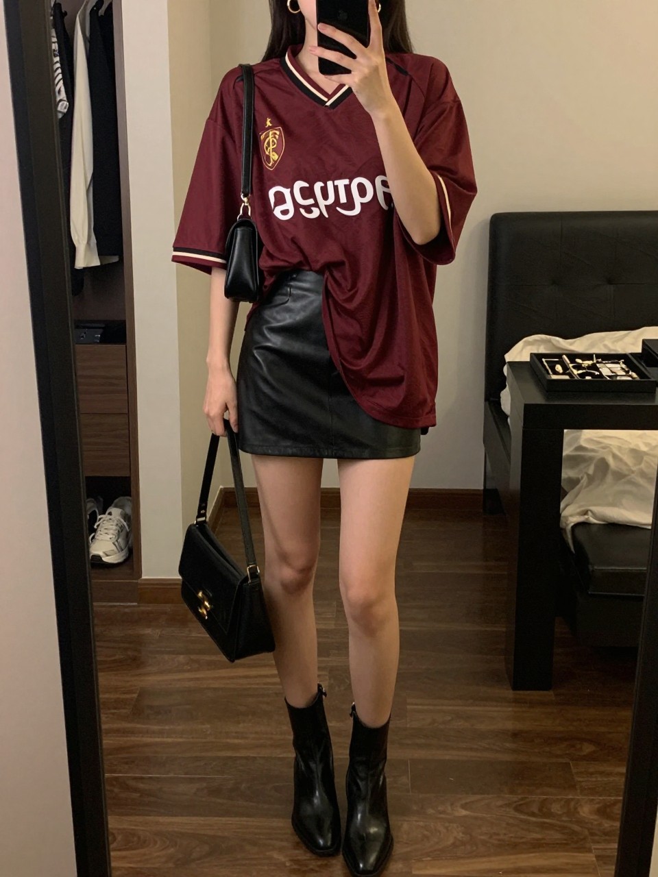mirror selfie of a woman wearing an oversized burgundy soccer jersey, black faux leather mini skirt, black ankle boots, black mini shoulder bag, and gold hoop earrings, clearly showing the same clothing items, colors, fabrics, and accessories listed in the Outfit Pieces section, holding a phone in front of a mirror, cropped at NECK so the face does not show, with a relaxed casual selfie pose, in a stylish city apartment, warm moody lighting, including an open closet with clothes visible and a jewelry tray on the vanity, latest iPhone photo quality.