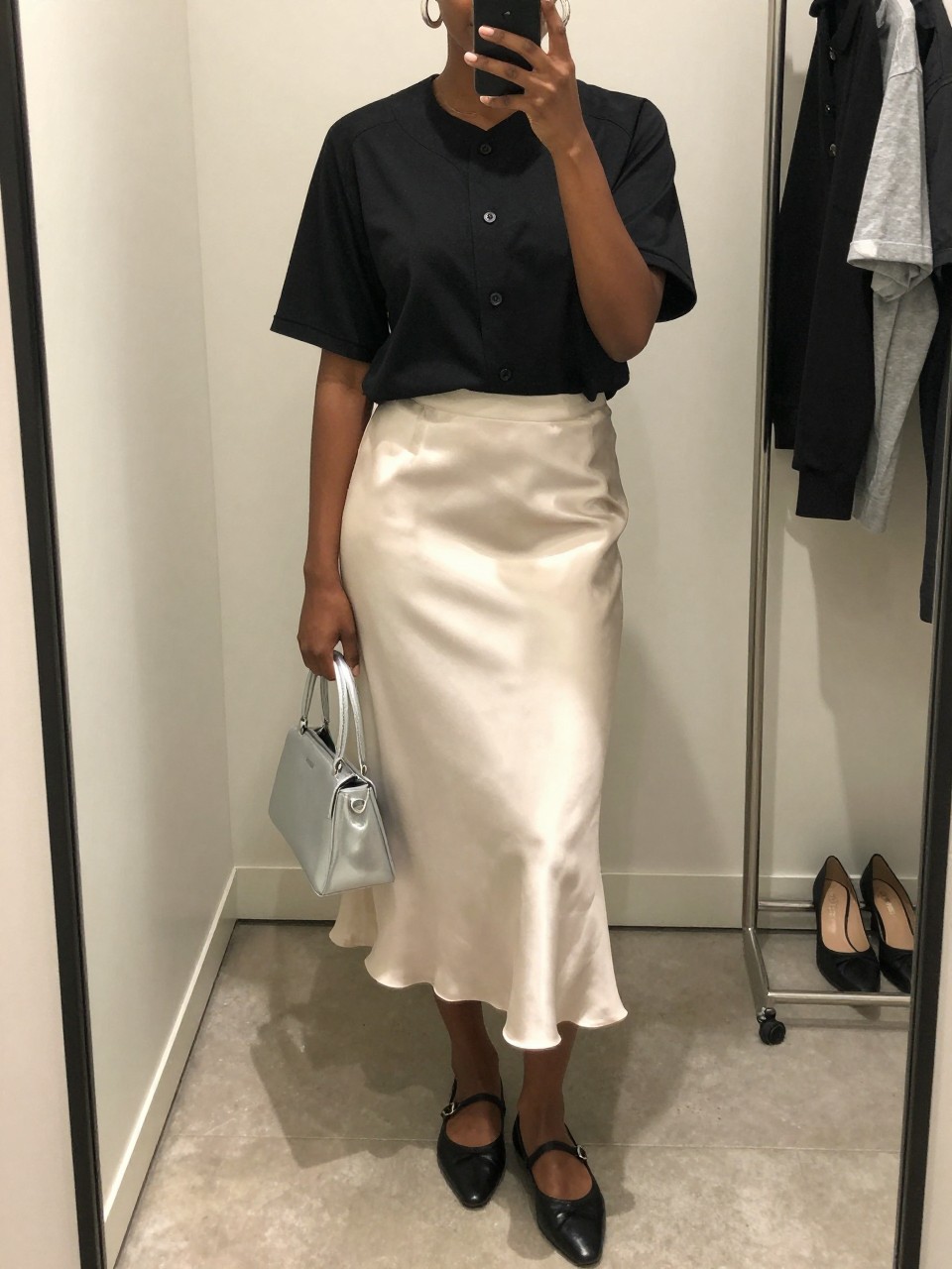 mirror selfie of a Black woman wearing a black button-front baseball jersey tucked into an ivory satin midi skirt, black pointed ballet flats, silver mini handbag, and chunky silver hoop earrings, clearly showing the same clothing items, colors, fabrics, and accessories listed in the Outfit Pieces section, holding a phone in front of a mirror, cropped at NECK so the face does not show, with a relaxed casual selfie pose, in a minimal dressing room, soft indoor lighting, including a clothing rack with garments behind and a pair of heels on the floor nearby, latest iPhone photo quality.