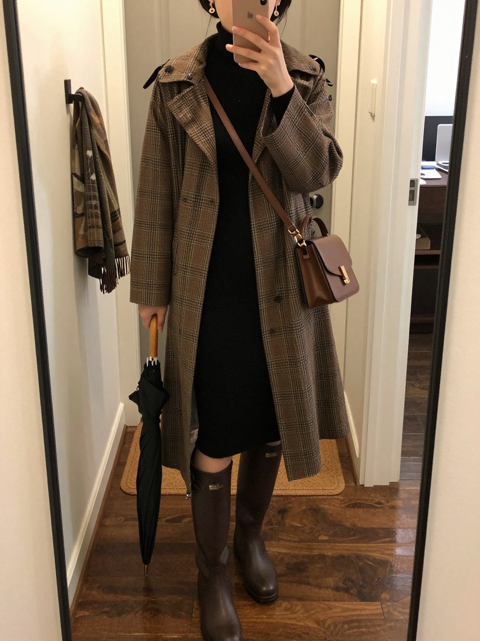 mirror selfie of a woman wearing the exact outfit described above, clearly showing a brown plaid water-resistant rain cape layered over a black fitted turtleneck midi dress, dark brown knee-high rain boots, a chocolate leather crossbody bag, a black umbrella in hand, and gold drop earrings, holding a phone in front of a mirror, cropped at NECK so the face does not show, with a relaxed casual selfie pose, in a warm apartment entryway mirror, soft rainy evening light, with a wet doormat near the door and a scarf hanging on a wall hook, latest iPhone photo quality.