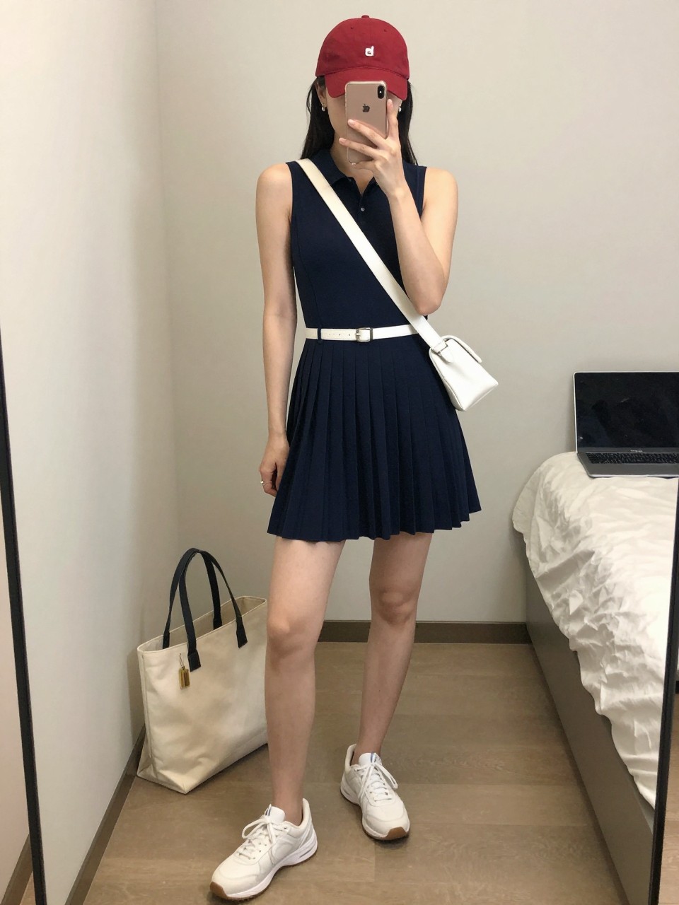 Image Prompt:
mirror selfie of a woman wearing a navy pleated sleeveless tennis dress, white athletic sneakers, a red cotton baseball cap, a white belt bag worn crossbody, and tiny silver huggie earrings, clearly showing the same clothing items, colors, fabrics, and accessories listed in the Outfit Pieces section, holding a phone in front of a mirror, cropped at NECK so the face does not show, with a relaxed casual selfie pose, in a minimal dressing room, soft indoor lighting, with a tote bag resting on the floor and a laptop or tablet on the bed, natural casual mirror selfie, not staged, latest iPhone photo quality.
