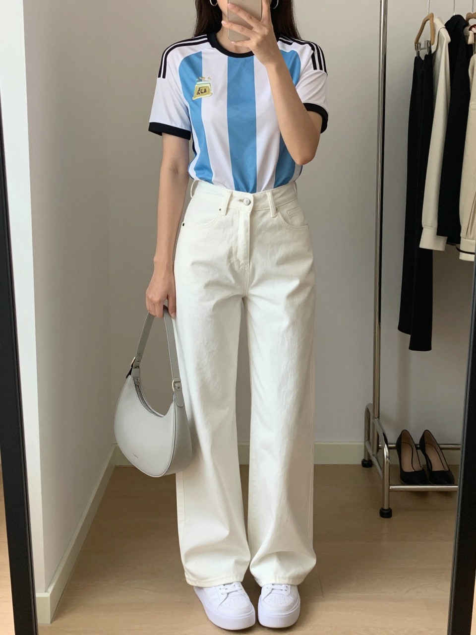 mirror selfie of a woman wearing a fitted sky blue and white Argentina soccer jersey tucked into white high-waisted wide-leg jeans, white platform sneakers, pale gray crescent shoulder bag, and silver hoop earrings, clearly showing the same clothing items, colors, fabrics, and accessories listed in the Outfit Pieces section, holding a phone in front of a mirror, cropped at NECK so the face does not show, with a relaxed casual selfie pose, in a minimal dressing room, soft indoor lighting, including a clothing rack with garments behind and a pair of heels on the floor nearby, latest iPhone photo quality.