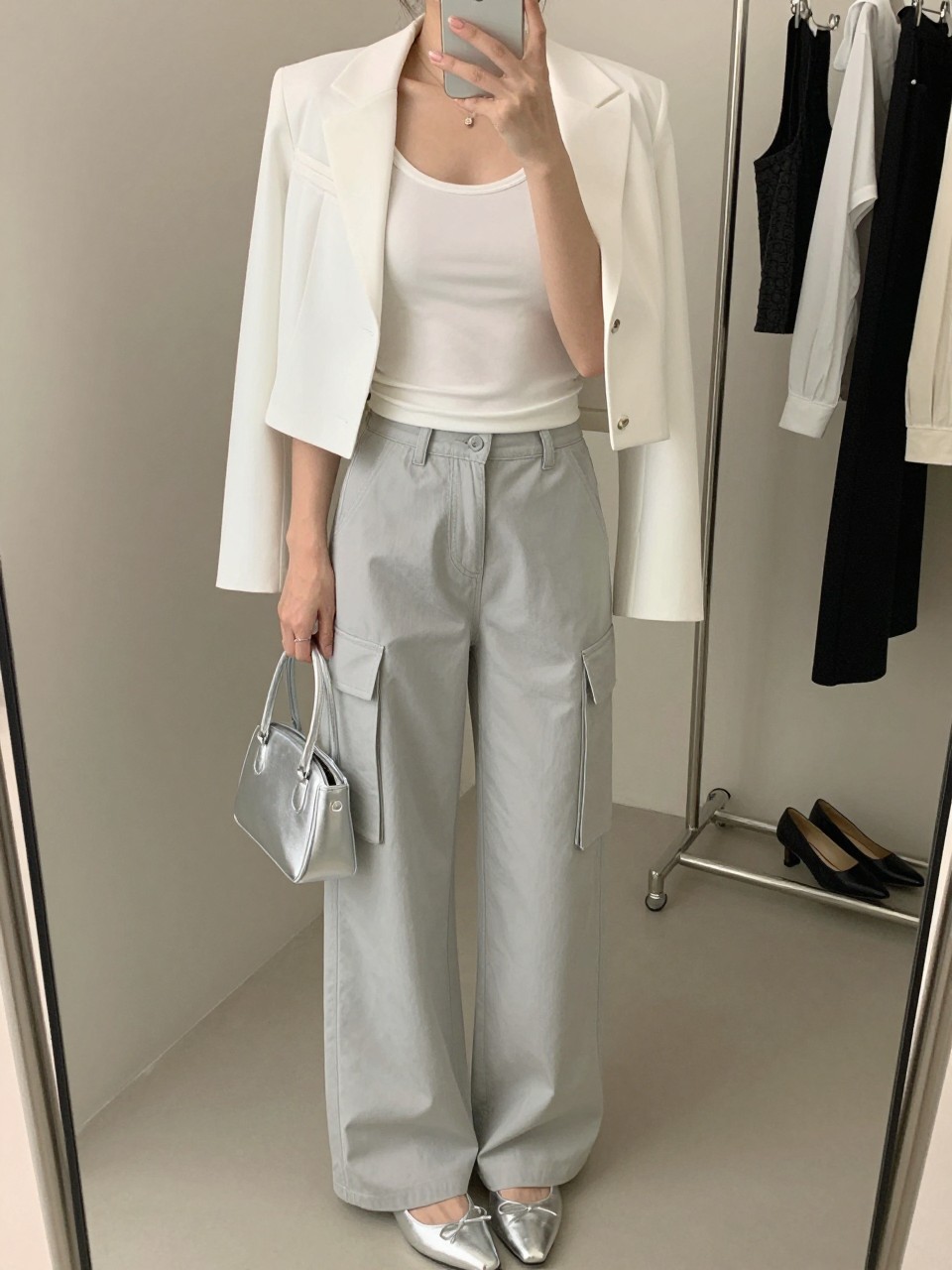 mirror selfie of a woman wearing light gray cargo pants, white fitted tank top, white cropped blazer, silver pointed flats, and silver mini handbag, clearly showing the same clothing items, colors, fabrics, and accessories listed in the Outfit Pieces section, holding a phone in front of a mirror, cropped at NECK so the face does not show, with a relaxed casual selfie pose, in a fashion studio mirror corner, soft indoor lighting, including a clothing rack with garments behind and a pair of heels on the floor nearby, latest iPhone photo quality.