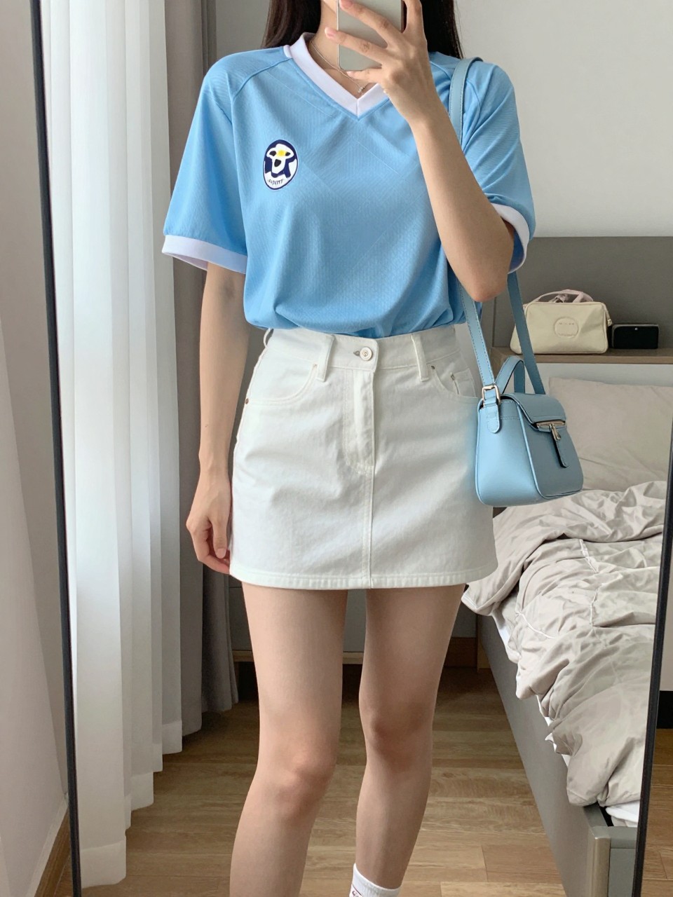 mirror selfie of a woman wearing a sky blue soccer jersey, white denim mini skirt, white low-top sneakers, pale blue mini shoulder bag, and delicate silver necklace, clearly showing the same clothing items, colors, fabrics, and accessories listed in the Outfit Pieces section, holding a phone in front of a mirror, cropped at NECK so the face does not show, with a relaxed casual selfie pose, in a cozy bedroom mirror setup, natural window light, including soft pillows on an unmade bed and a cosmetic bag on the counter, latest iPhone photo quality.