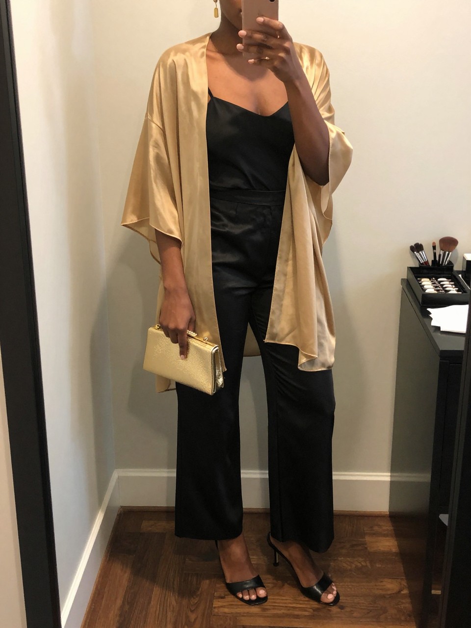 mirror selfie of a Black woman wearing a soft gold sheer poncho layered over a black fitted camisole, black satin wide-leg pants, black heeled mules, gold mini clutch, and gold drop earrings, clearly showing the same clothing items, colors, fabrics, and accessories listed in the Outfit Pieces section, holding a phone in front of a mirror, cropped at NECK so the face does not show, with a relaxed casual selfie pose, in a fashion studio mirror corner, warm moody lighting, including a makeup brush on the counter and a jewelry tray on the vanity, latest iPhone photo quality.