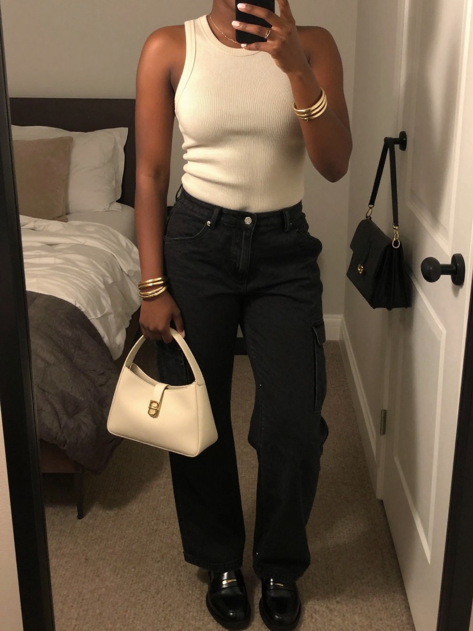 mirror selfie of a Black woman wearing black cargo jeans, cream fitted knit tank top, black leather loafers, cream structured mini bag, and gold layered bracelets, clearly showing the same clothing items, colors, fabrics, and accessories listed in the Outfit Pieces section, holding a phone in front of a mirror, cropped at NECK so the face does not show, with a relaxed casual selfie pose, in a cozy bedroom mirror setup, warm moody lighting, including soft pillows on an unmade bed and a handbag hanging on a door handle, latest iPhone photo quality.