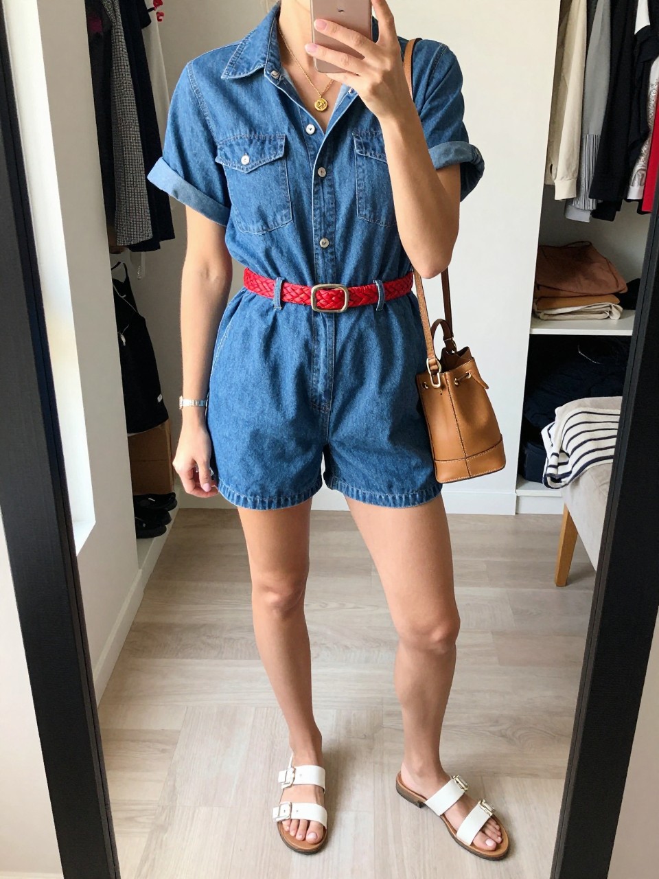 Image Prompt:
mirror selfie of a woman wearing a blue denim short-sleeve romper, a red woven waist belt, white flat sandals, a small tan bucket bag, and a gold charm necklace, clearly showing the same clothing items, colors, fabrics, and accessories listed in the Outfit Pieces section, holding a phone in front of a mirror, cropped at NECK so the face does not show, with a relaxed casual selfie pose, in a walk-in closet mirror, natural window light, with an open closet with clothes visible and a folded blanket on a chair, natural casual mirror selfie, not staged, latest iPhone photo quality.