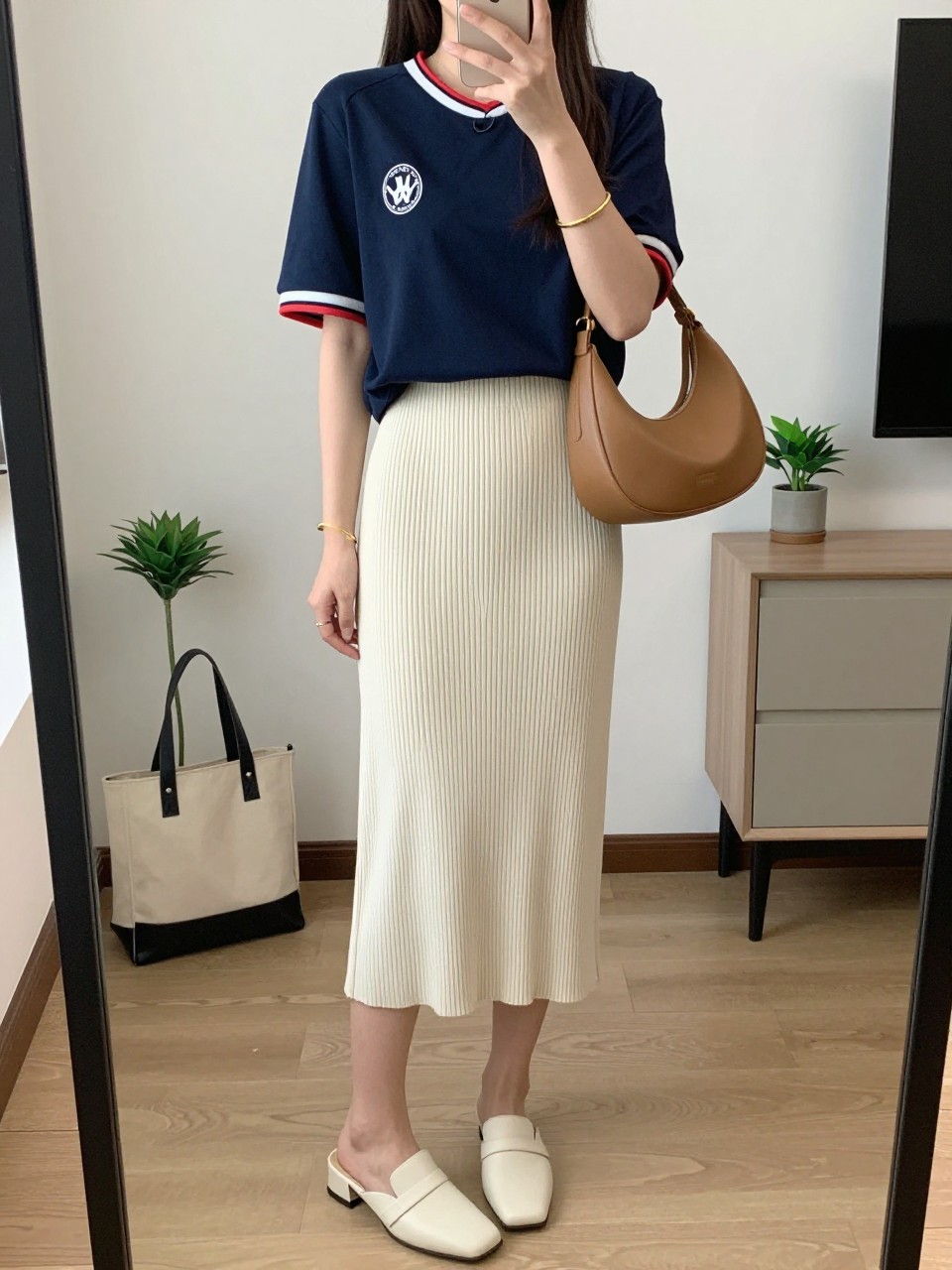 mirror selfie of a woman wearing a navy soccer jersey tucked into a cream ribbed knit midi skirt, cream heeled mules, tan crescent shoulder bag, and gold bangle bracelet, clearly showing the same clothing items, colors, fabrics, and accessories listed in the Outfit Pieces section, holding a phone in front of a mirror, cropped at NECK so the face does not show, with a relaxed casual selfie pose, in a stylish city apartment, soft indoor lighting, including a small plant on a dresser and a tote bag resting on the floor, latest iPhone photo quality.