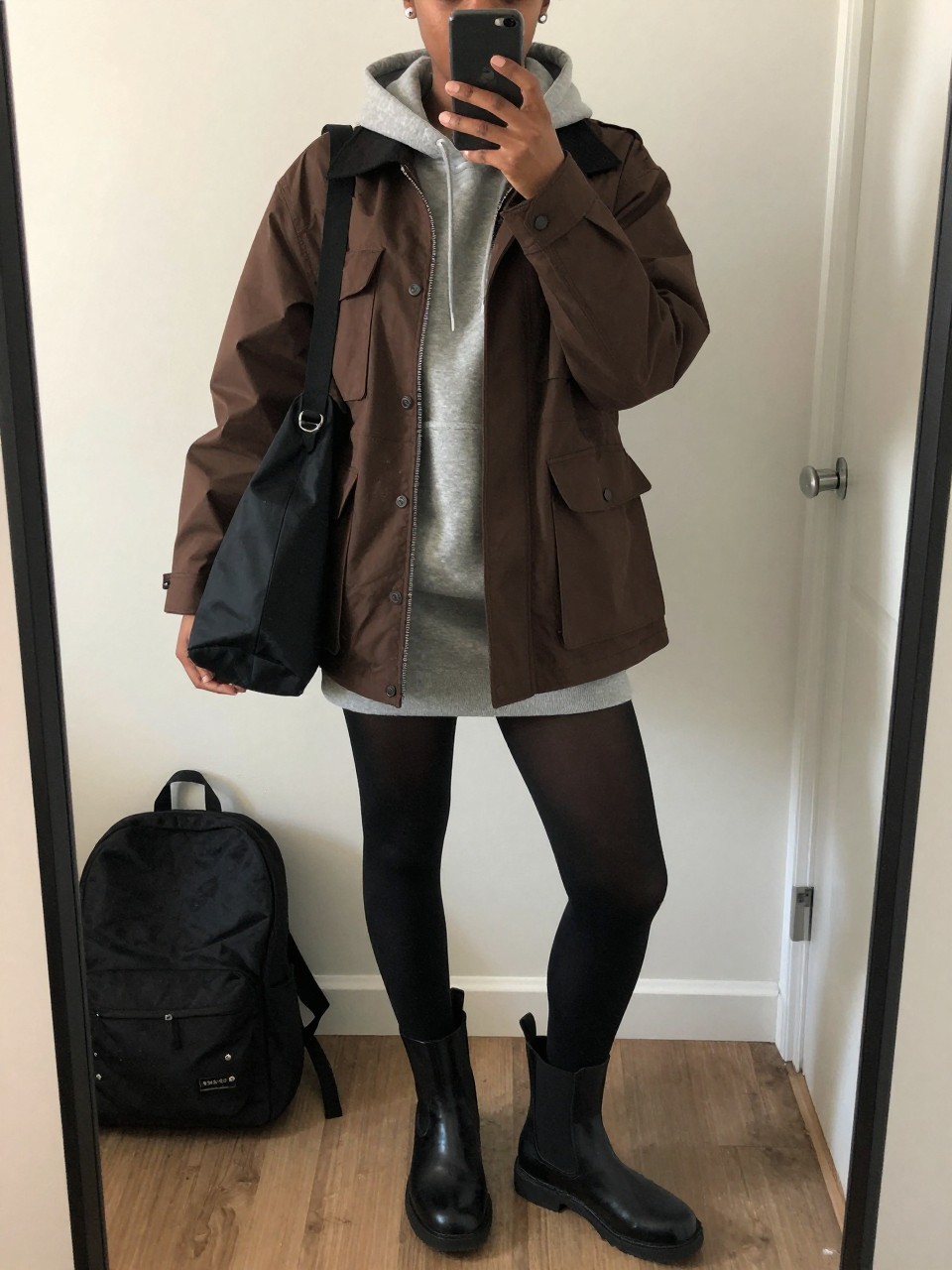 mirror selfie of a Black woman wearing the exact outfit described above, clearly showing a brown water-resistant utility jacket layered over a gray hoodie mini dress, black opaque tights, black waterproof Chelsea boots, a black nylon shoulder bag, and silver stud earrings, holding a phone in front of a mirror, cropped at NECK so the face does not show, with a relaxed casual selfie pose, in a casual apartment mirror, cloudy morning light, with a backpack leaning against the wall and a damp umbrella near the door, latest iPhone photo quality.