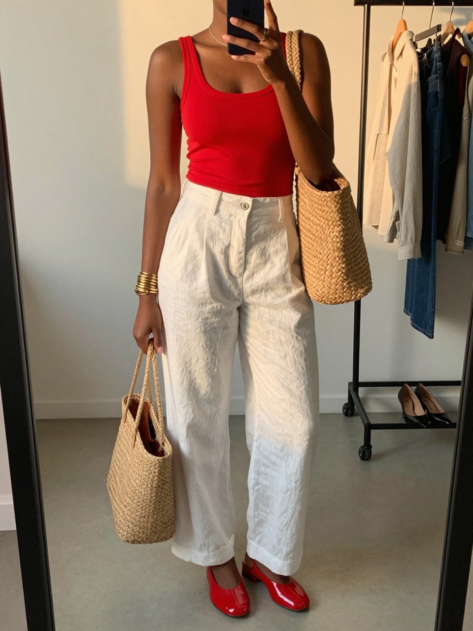 mirror selfie of a Black woman wearing white high-waisted linen pants, a red fitted tank top, glossy red jelly shoes, tan woven tote bag, and gold layered bracelets, clearly showing the same clothing items, colors, fabrics, and accessories listed in the Outfit Pieces section, holding a phone in front of a mirror, cropped at NECK so the face does not show, with a relaxed casual selfie pose, in a minimal dressing room, evening golden hour, including a clothing rack with garments behind and a pair of heels on the floor nearby, latest iPhone photo quality.