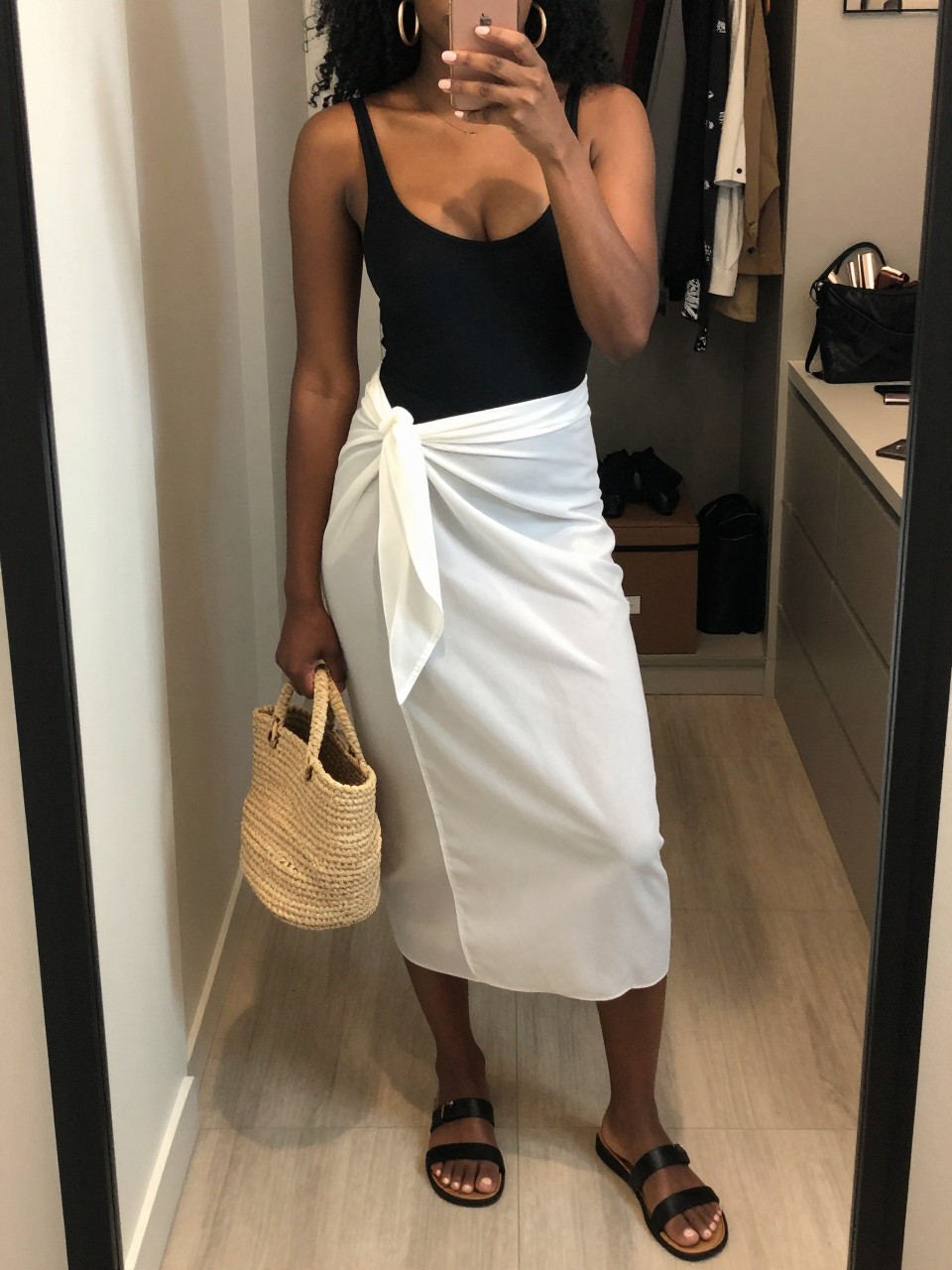 mirror selfie of a Black woman wearing a black one-piece swimsuit, white lightweight sarong skirt tied at the waist, black flat sandals, straw bucket bag, and chunky gold hoop earrings, clearly showing the same clothing items, colors, fabrics, and accessories listed in the Outfit Pieces section, holding a phone in front of a mirror, cropped at NECK so the face does not show, with a relaxed casual selfie pose, in a walk-in closet mirror, soft indoor lighting, including an open closet with clothes visible and a cosmetic bag on the counter, latest iPhone photo quality.