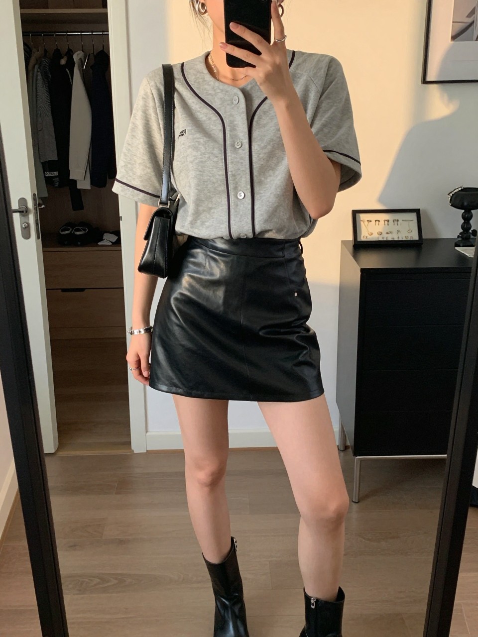 mirror selfie of a woman wearing a gray button-front baseball jersey, black faux leather mini skirt, black ankle boots, black mini shoulder bag, and silver hoop earrings, clearly showing the same clothing items, colors, fabrics, and accessories listed in the Outfit Pieces section, holding a phone in front of a mirror, cropped at NECK so the face does not show, with a relaxed casual selfie pose, in a stylish city apartment, evening golden hour, including an open closet with clothes visible and a jewelry tray on the vanity, latest iPhone photo quality.
