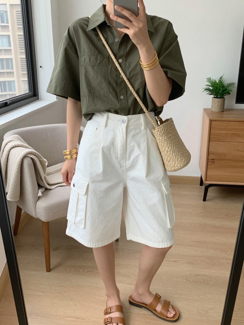 mirror selfie of a woman wearing white baggy cargo shorts, olive green cropped button-up shirt, tan leather slide sandals, woven beige crossbody bag, and gold layered bracelets, clearly showing the same clothing items, colors, fabrics, and accessories listed in the Outfit Pieces section, holding a phone in front of a mirror, cropped at NECK so the face does not show, with a relaxed casual selfie pose, in a stylish city apartment, natural window light, including a folded blanket on a chair and a small plant on a dresser, latest iPhone photo quality.