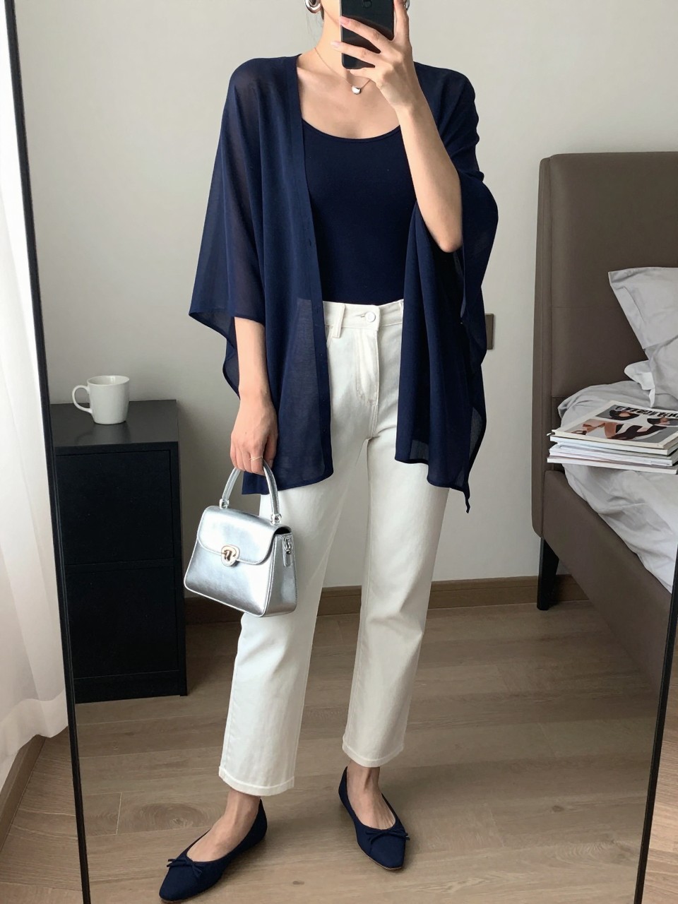 mirror selfie of a woman wearing a navy sheer poncho layered over a navy fitted tank top, white straight-leg jeans, navy pointed ballet flats, silver mini handbag, and silver hoop earrings, clearly showing the same clothing items, colors, fabrics, and accessories listed in the Outfit Pieces section, holding a phone in front of a mirror, cropped at NECK so the face does not show, with a relaxed casual selfie pose, in a stylish city apartment, soft indoor lighting, including a coffee mug on a bedside table and a stack of fashion magazines on a side table, latest iPhone photo quality.