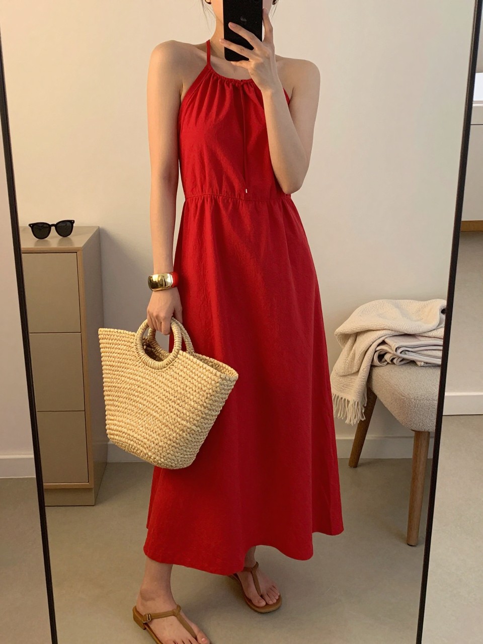 Image Prompt:
mirror selfie of a woman wearing a red cotton halter maxi dress, tan flat thong sandals, a large woven straw tote, a chunky gold bangle, and a sleek low bun, clearly showing the same clothing items, colors, fabrics, and accessories listed in the Outfit Pieces section, holding a phone in front of a mirror, cropped at NECK so the face does not show, with a relaxed casual selfie pose, in a minimal dressing room, evening golden hour, with sunglasses placed on a dresser and a folded blanket on a chair, natural casual mirror selfie, not staged, latest iPhone photo quality.