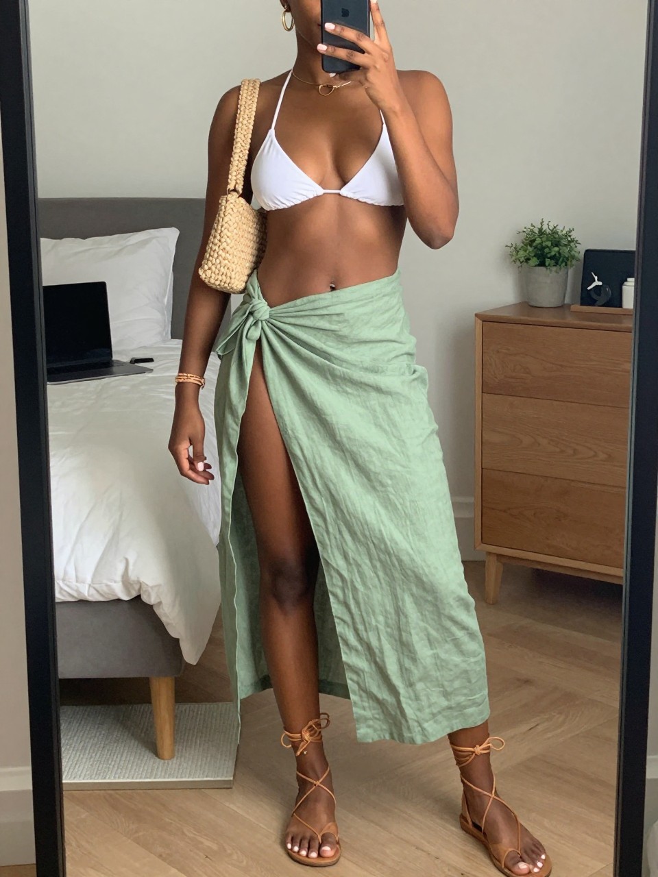 mirror selfie of a Black woman wearing a white bikini top, sage green linen wrap skirt, tan lace-up flat sandals, woven mini shoulder bag, and gold hoop earrings, clearly showing the same clothing items, colors, fabrics, and accessories listed in the Outfit Pieces section, holding a phone in front of a mirror, cropped at NECK so the face does not show, with a relaxed casual selfie pose, in a stylish city apartment, soft indoor lighting, including a laptop on the bed and a small plant on a dresser, latest iPhone photo quality.