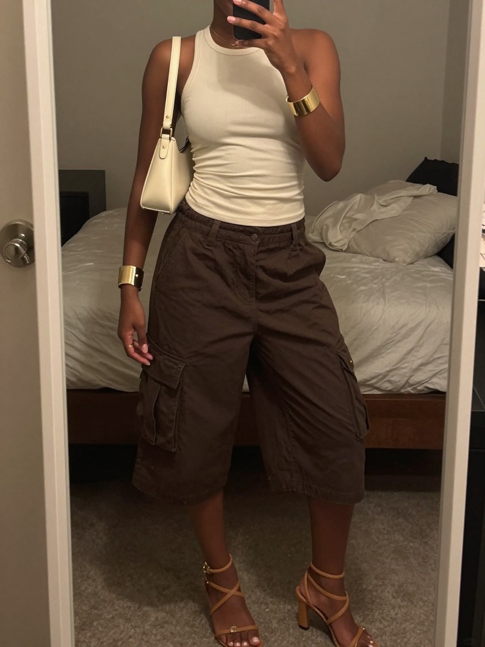 mirror selfie of a Black woman wearing chocolate brown baggy cargo shorts, cream fitted halter top, tan strappy heeled sandals, cream mini shoulder bag, and gold cuff bracelet, clearly showing the same clothing items, colors, fabrics, and accessories listed in the Outfit Pieces section, holding a phone in front of a mirror, cropped at NECK so the face does not show, with a relaxed casual selfie pose, in a cozy bedroom mirror setup, warm moody lighting, including soft pillows on an unmade bed and a handbag hanging on a door handle, latest iPhone photo quality.