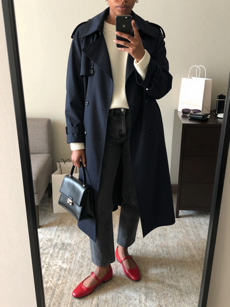 mirror selfie of a Black woman wearing the exact outfit described above, clearly showing a black trench coat layered over a cream ribbed sweater, dark gray straight-leg jeans, red leather Mary Jane flats, a black mini handbag, and pearl stud earrings, holding a phone in front of a mirror, cropped at NECK so the face does not show, with a relaxed casual selfie pose, in a stylish Tokyo hotel mirror, soft natural light, with a small shopping bag near the dresser and sunglasses beside a travel wallet, latest iPhone photo quality.