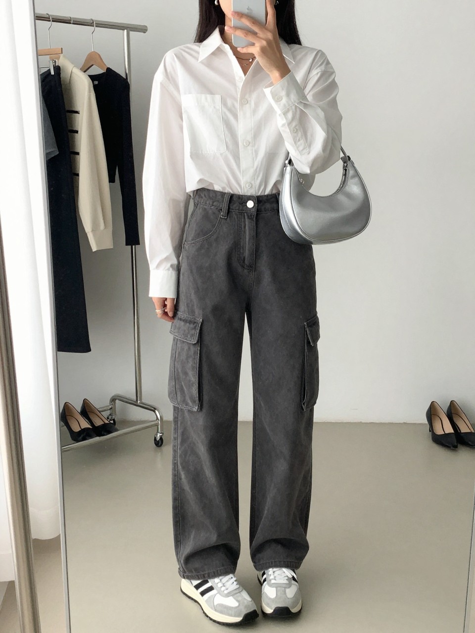 mirror selfie of a woman wearing gray cargo jeans, cropped white button-down shirt, white and silver sneakers, silver crescent shoulder bag, and small silver huggie earrings, clearly showing the same clothing items, colors, fabrics, and accessories listed in the Outfit Pieces section, holding a phone in front of a mirror, cropped at NECK so the face does not show, with a relaxed casual selfie pose, in a minimal dressing room, natural window light, including a clothing rack with garments behind and a pair of heels on the floor nearby, latest iPhone photo quality.