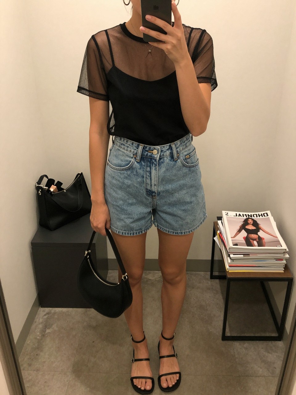 Outfit Image Prompt:
mirror selfie of a woman wearing a black sheer mesh short-sleeve top layered over a black fitted camisole, light-wash high-waisted denim shorts, black strappy flat sandals, small black crescent bag, and silver hoop earrings, holding a phone in front of a mirror, cropped at NECK so the face does not show, relaxed downtown NYC selfie pose, in a minimal dressing room, warm moody lighting, with a makeup bag on the counter and a stack of fashion magazines on a side table, realistic latest iPhone photo quality, casual summer street style.