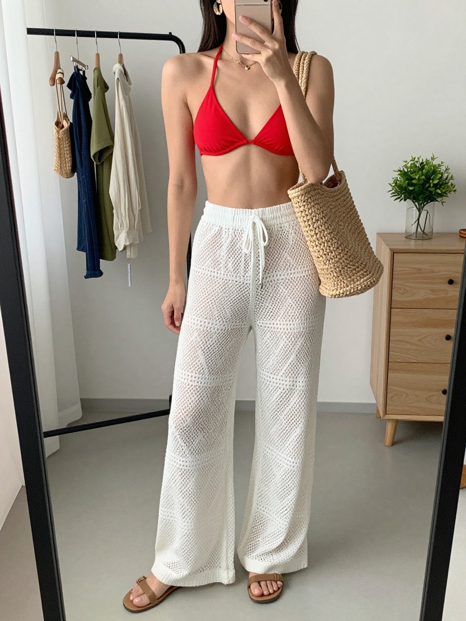 mirror selfie of a woman wearing a red bikini top, white crochet wide-leg pants, tan flat sandals, raffia beach tote, and gold shell earrings, clearly showing the same clothing items, colors, fabrics, and accessories listed in the Outfit Pieces section, holding a phone in front of a mirror, cropped at NECK so the face does not show, with a relaxed casual selfie pose, in a fashion studio mirror corner, natural window light, including a clothing rack with garments behind and a small plant on a dresser, latest iPhone photo quality.
