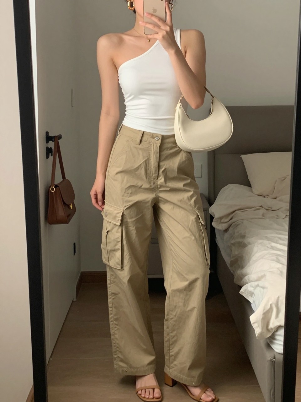 mirror selfie of a woman wearing khaki cargo pants, fitted white one-shoulder top, tan heeled sandals, cream crescent shoulder bag, and chunky gold hoop earrings, clearly showing the same clothing items, colors, fabrics, and accessories listed in the Outfit Pieces section, holding a phone in front of a mirror, cropped at NECK so the face does not show, with a relaxed casual selfie pose, in a cozy bedroom mirror setup, soft indoor lighting, including soft pillows on an unmade bed and a handbag hanging on a door handle, latest iPhone photo quality.
