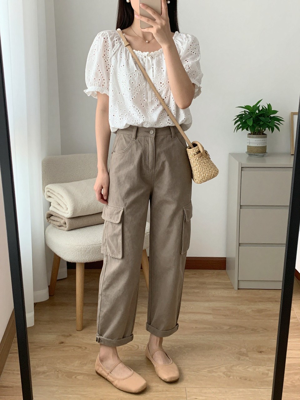 mirror selfie of a woman wearing taupe cargo pants, white eyelet short-sleeve blouse, nude ballet flats, woven beige crossbody bag, and pearl stud earrings, clearly showing the same clothing items, colors, fabrics, and accessories listed in the Outfit Pieces section, holding a phone in front of a mirror, cropped at NECK so the face does not show, with a relaxed casual selfie pose, in a feminine-themed bedroom, natural window light, including a folded blanket on a chair and a small plant on a dresser, latest iPhone photo quality.