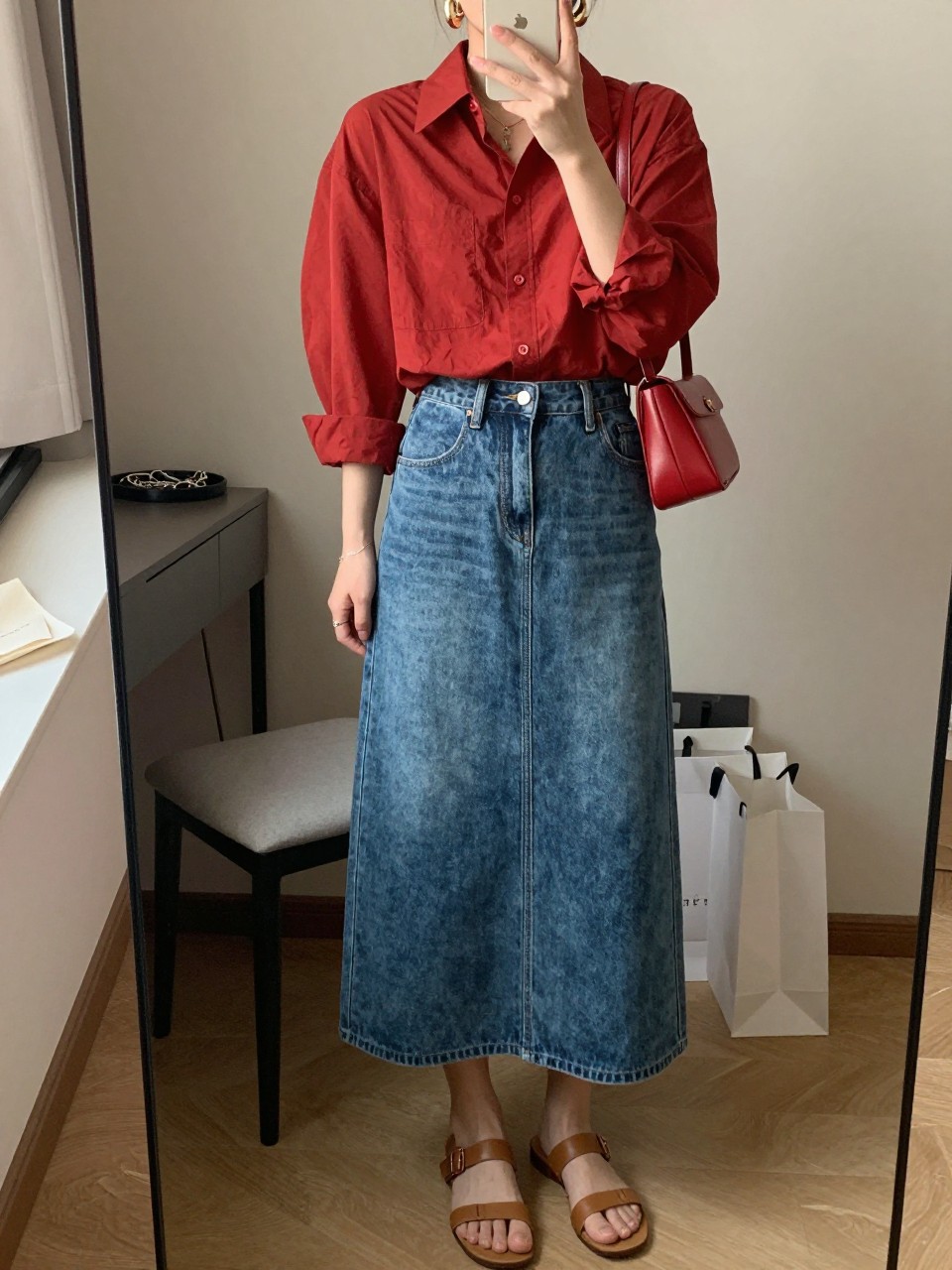 mirror selfie of a woman wearing a red oversized button down shirt tucked into a medium blue denim maxi skirt with front slit, tan leather flat sandals, red mini shoulder bag, and chunky gold hoop earrings, clearly showing the same clothing items, colors, fabrics, and accessories listed in the Outfit Pieces section, holding a phone in front of a mirror, cropped at NECK so the face does not show, with a relaxed casual selfie pose, in a cozy bedroom mirror setup, warm moody lighting, including a jewelry tray on the vanity and shopping bags beside a chair, latest iPhone photo quality.