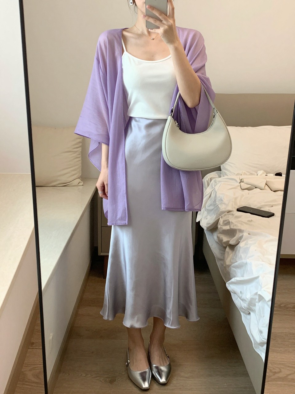 mirror selfie of a woman wearing a lavender sheer poncho layered over a white fitted camisole, light gray satin slip skirt, silver pointed flats, pale gray crescent shoulder bag, and small silver huggie earrings, clearly showing the same clothing items, colors, fabrics, and accessories listed in the Outfit Pieces section, holding a phone in front of a mirror, cropped at NECK so the face does not show, with a relaxed casual selfie pose, in a cozy bedroom mirror setup, soft indoor lighting, including soft pillows on an unmade bed and a cosmetic bag on the counter, latest iPhone photo quality.