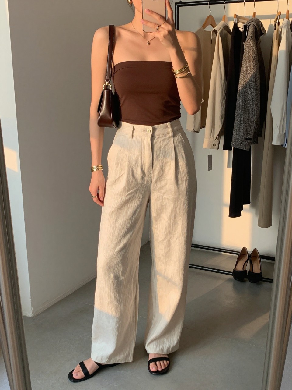 Outfit Image Prompt:
mirror selfie of a woman wearing a brown fitted tube top, cream high-waisted linen trousers, black flat sandals, small chocolate brown shoulder bag, and gold layered bracelets, holding a phone in front of a mirror, cropped at NECK so the face does not show, relaxed polished city selfie pose, in a fashion studio mirror corner, evening golden hour lighting, with a clothing rack with garments behind and a pair of heels on the floor nearby, realistic latest iPhone photo quality, soft summer NYC styling.