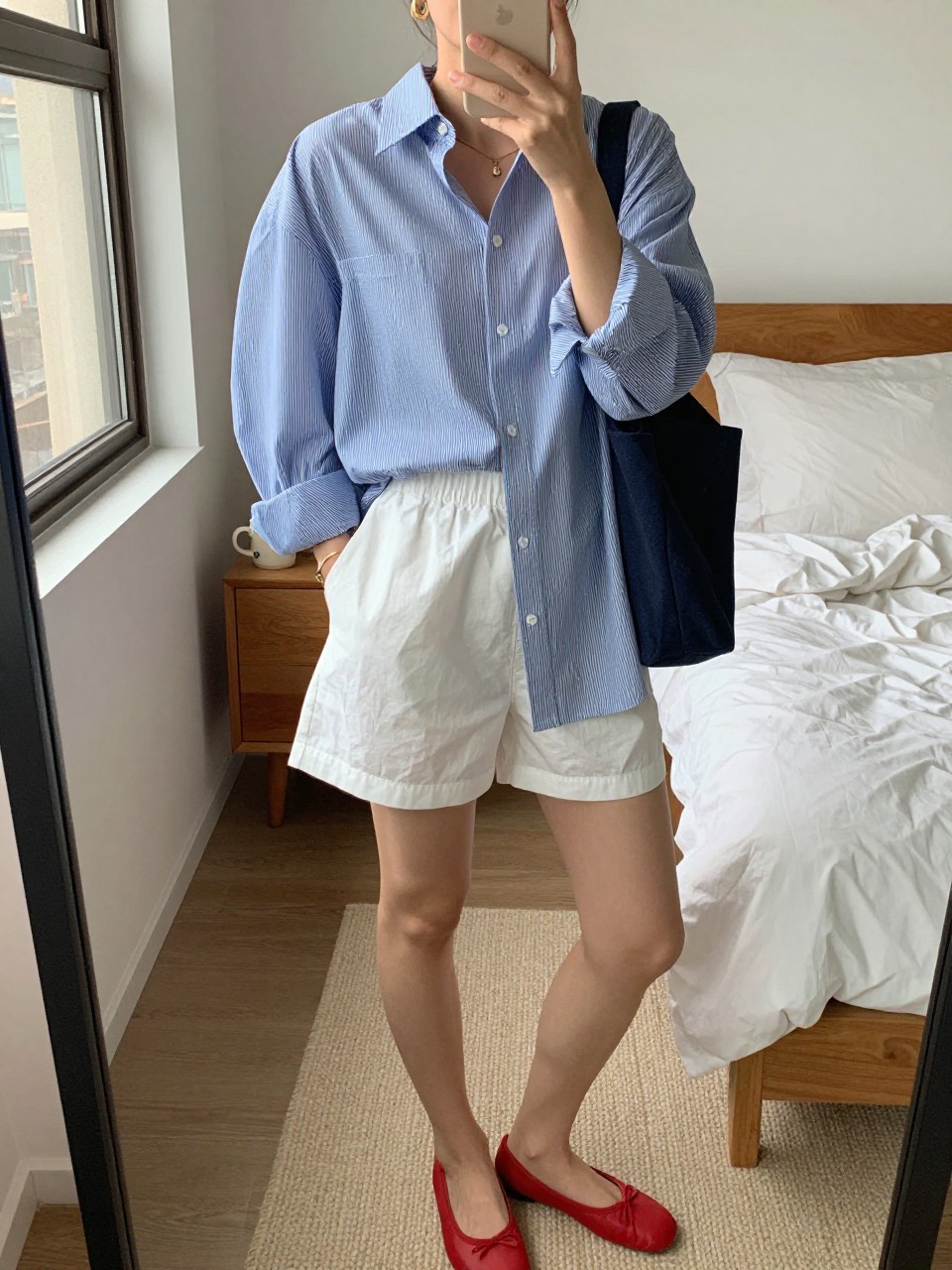 Outfit Image Prompt:
mirror selfie of a woman wearing an oversized blue and white striped cotton button-down shirt, white cotton boxer-style shorts, red ballet flats, small navy canvas tote bag, and gold huggie earrings, holding a phone in front of a mirror, cropped at NECK so the face does not show, relaxed NYC summer selfie pose, in a cozy bedroom mirror setup, natural window light, with a coffee mug on a bedside table and soft pillows on an unmade bed, realistic latest iPhone photo quality, casual lived-in styling.