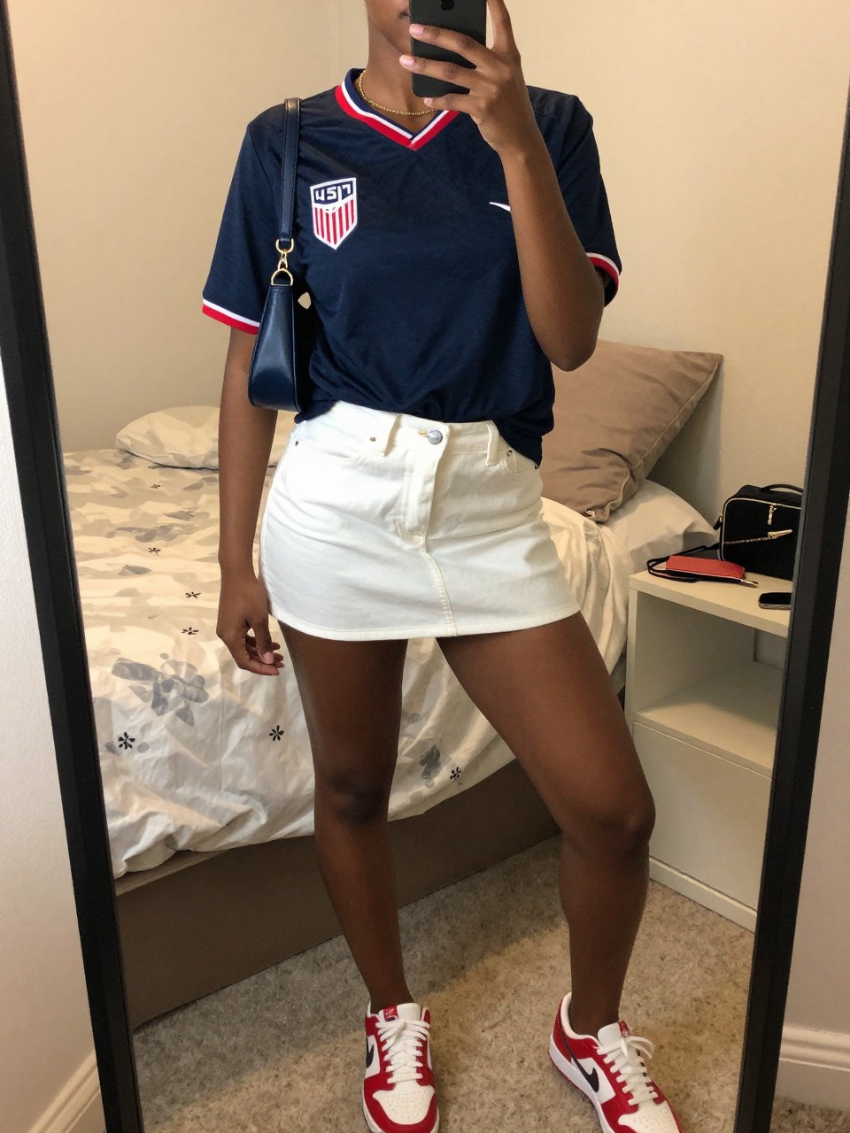 mirror selfie of a Black woman wearing a navy USA soccer jersey, white denim mini skirt, red and white sneakers, navy mini shoulder bag, and slim gold chain necklace, clearly showing the same clothing items, colors, fabrics, and accessories listed in the Outfit Pieces section, holding a phone in front of a mirror, cropped at NECK so the face does not show, with a relaxed casual selfie pose, in a cozy bedroom mirror setup, evening golden hour, including soft pillows on an unmade bed and a cosmetic bag on the counter, latest iPhone photo quality.