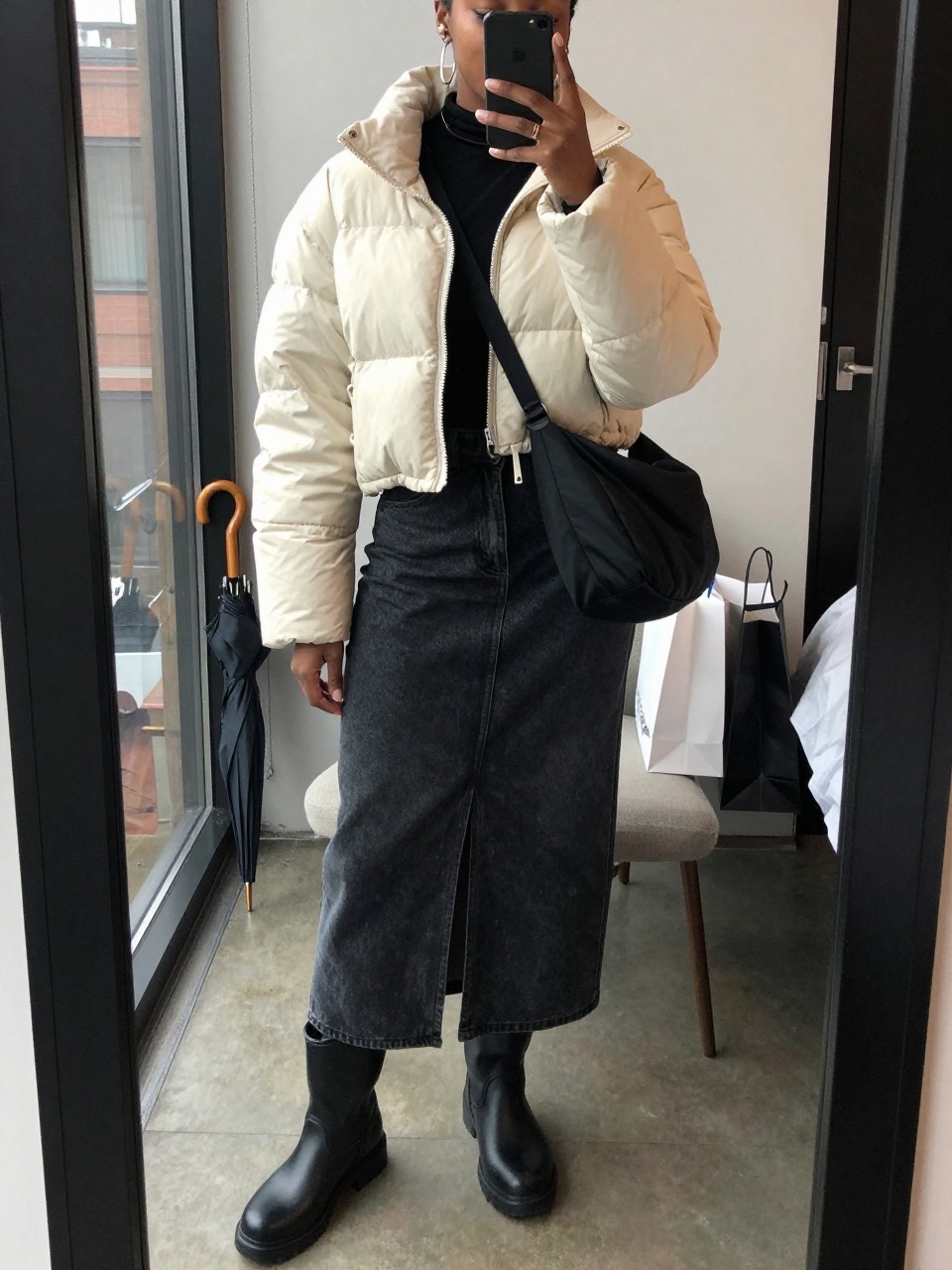 mirror selfie of a Black woman wearing the exact outfit described above, clearly showing a cream cropped water-resistant puffer jacket layered over a fitted black long-sleeve top, dark denim maxi skirt with a front slit, black lug-sole rain boots, a black nylon crossbody bag, and silver hoop earrings, holding a phone in front of a mirror, cropped at NECK so the face does not show, with a relaxed casual selfie pose, in a stylish city apartment mirror, soft cloudy window light, with shopping bags beside a chair and a wet umbrella near the door, latest iPhone photo quality.