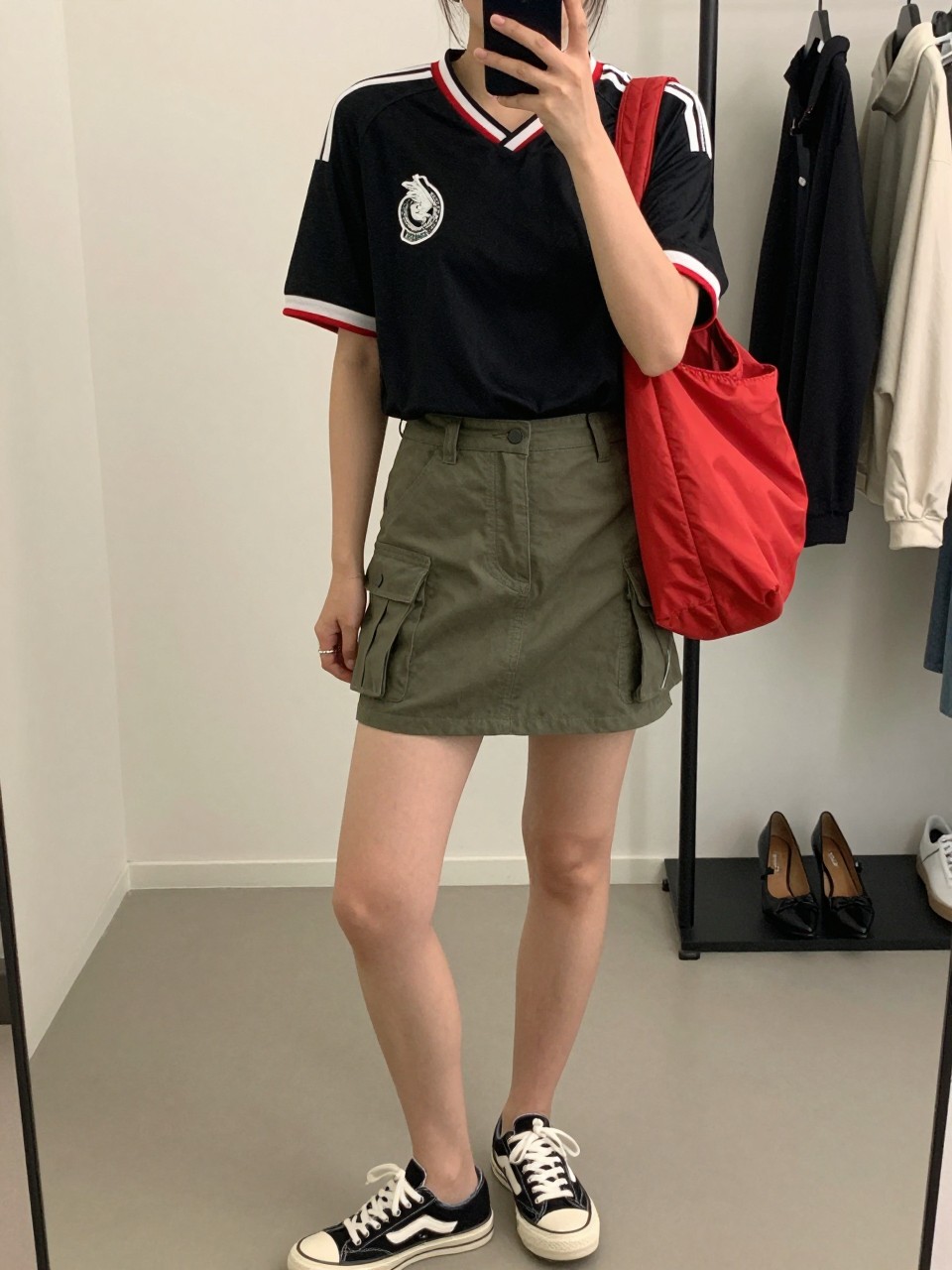 mirror selfie of a woman wearing a black Mexico soccer jersey, olive green cargo mini skirt, black and white sneakers, red nylon shoulder bag, and silver hoop earrings, clearly showing the same clothing items, colors, fabrics, and accessories listed in the Outfit Pieces section, holding a phone in front of a mirror, cropped at NECK so the face does not show, with a relaxed casual selfie pose, in a minimal dressing room, soft indoor lighting, including a clothing rack with garments behind and a pair of heels on the floor nearby, latest iPhone photo quality.